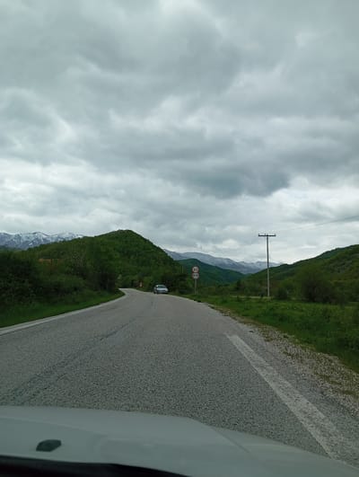 The image depicts a road curving through a mountainous landscape under an overcast sky, potentially located near Melivoia, Greece. A white car is partially visible in the bottom right corner, suggesting the photo was taken from inside a vehicle. The road is flanked by green vegetation and mountains. In the distance, snow-capped peaks are visible through a break in the clouds. A utility pole and wires run parallel to the road. A speed limit sign is also visible. Another vehicle is further down the road.