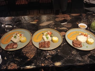 Three plates of food are presented on a dark, granite-patterned tabletop. Each plate contains what appears to be a smear of orange puree, a portion of rice, a piece of corn on the cob, and a cut of meat. One plate includes peas and some cooked vegetables. A small bowl of sauce is visible near the right plate, along with a jar of green liquid. In the background, there are chairs and standing people. There is no evidence to link the image to Nwanaeanoka, Botswana. The description is based solely on the visual details present in the image.