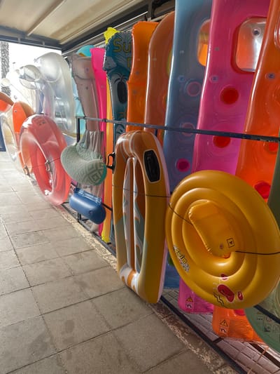 The image displays a large selection of inflatable water toys and accessories arranged on a multi-tiered metal rack, positioned outdoors in L'estartit, Spain. The items are brightly colored, including shades of orange, pink, blue, yellow, clear, and white, with some featuring patterns or text such as "SWIM" and "SUMMER." Visible objects include numerous inflatable rings of varying diameters, several long inflatable mattresses or lounges, a light blue patterned inflatable neck pillow, and a small, dark blue cylindrical inflatable. These items are suspended from horizontal black metal bars, forming a display against a background of additional inflatables. The ground consists of light grey square paver tiles. An overhead awning or roof structure is visible above the display. In the blurred background, partial views of distant palm trees or coastal vegetation are discernable under an overcast sky. No individuals are present in the scene. The context suggests an outdoor retail area, likely a beach or resort shop.