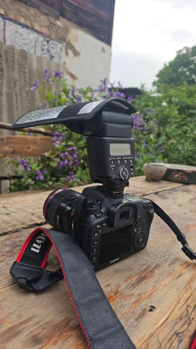The image shows a Canon DSLR camera with a flash attached, resting on a weathered wooden surface. A black camera strap with red trim is visible, bearing the "Canon" logo. In the background, there are out-of-focus purple flowers and greenery, suggesting an outdoor setting. The lighting appears diffused, indicating an overcast day. The overall scene is a close-up shot of photography equipment, possibly staged for a demonstration or simply left on a table outdoors. There are no people or animals visible. The visible text includes "Canon", "Canon Lens", and various camera function labels such as "MENU", "INFO", "MODE", "ZOOM", "PILOT", "ON", "OFF", "RATE", and "AF-ON". The location context provided is Панфилово, Kazakhstan, which adds a geographical detail to the scene.