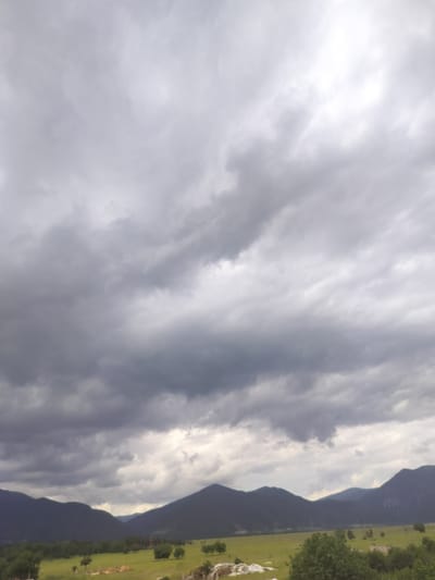 The image depicts a cloudy sky over a rural landscape in Gorno Sahrane, Bulgaria. Dark, ominous clouds dominate the upper portion of the frame, suggesting an impending storm. Below the sky, rolling hills and mountains form the horizon. The midground features a lush green field dotted with trees. In the foreground, the terrain is more varied, with patches of grass, some exposed soil, and a few scattered structures or building materials. The overall atmosphere is overcast and somber. There are no people or animals visible in the scene.