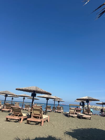 The image shows a beach in Zygi, Cyprus, featuring a row of straw umbrellas and beach chairs aligned along the shore. The scene is set on a sunny day with a clear blue sky. The beach appears to be sandy, with dark-colored sand. There are a few people visible in the distance, relaxing on the beach chairs under the umbrellas. The sea is calm, with a few small boats or ships visible in the horizon. A small breakwater is visible in the distance.