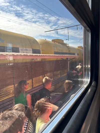 The image depicts a scene viewed from inside a train window, looking out onto an adjacent railway track and platform, ostensibly in the city of Vyatskiye Polyany, Russia. In the foreground, the dark frame of the train window is prominent, showing reflections of passengers inside the viewing train, including a woman in a green top, a man with light hair, and other partially visible individuals.

Beyond the window, a long freight train with multiple yellow tanker cars is stationary on a parallel track. One tanker car clearly displays "МЕТАНОЛ «Х»" (METHANOL "X") and "ЩЕКИНАЗОТ" (SHCHEKINAZOT) in Cyrillic lettering, indicating its contents and likely a chemical company. The sky above is clear blue with scattered white clouds, and overhead electrical lines for trains are visible.

Between the stationary freight train and the vantage point, a railway platform is visible. It features a reddish corrugated metal roof supported by posts, below which is a metal mesh fence with a hexagonal pattern. Several wooden benches are present on the platform side of the fence. A few people are visible on the platform: an adult individual wearing a light-colored top is seen from behind, walking away from the camera's perspective, while other figures are less distinct, also moving on the platform. The scene is illuminated by bright daylight, casting strong shadows and highlights.