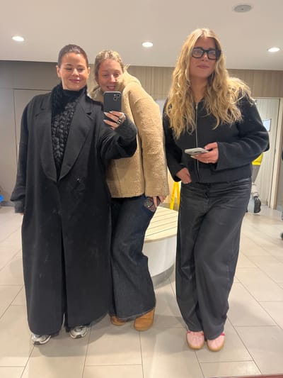 Three women pose for a selfie in what appears to be a public space with tiled flooring. The woman in the foreground wears a long black coat over a knit top. The woman in the middle has on a tan coat and denim pants. The third woman, at the top, sports a black zip-up jacket and matching pants. All three appear to be smiling and looking at the camera, with one woman holding a smartphone.