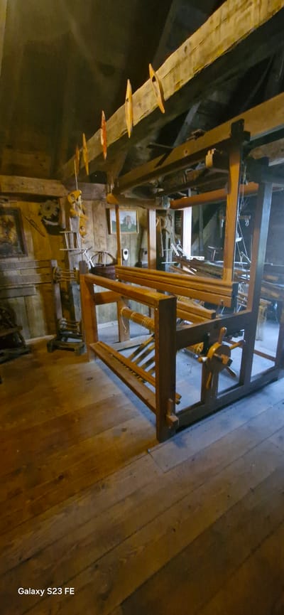The image shows the interior of a historical building, possibly a museum or a preserved workshop, in Pakruojis, Lithuania. The scene is dominated by a large, wooden loom, the central subject of the photograph. The loom is constructed from rough-hewn wooden beams and appears to be a traditional, manual textile-making device. Various wooden components, including rollers, beams, and parts of the weaving mechanism, are visible.

In the background, the walls are made of wood and brick, contributing to the rustic, historical ambiance. There are framed pictures or paintings hanging on the walls, depicting what appear to be portraits or scenes from rural life. Several wooden tools and implements are also present, scattered around the loom and on shelves, suggesting the space was used for craft or production. The floor is made of wooden planks, worn with age, and illuminated by natural light filtering in from unseen sources, creating a warm, dim atmosphere. The overall impression is one of a preserved historical setting, showcasing traditional craftsmanship.