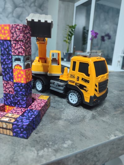 This media file features a toy excavation truck and a stack of colorful, pixelated blocks, likely resembling Minecraft-style game blocks. The truck is bright yellow with black accents and some text and graphics on its side. The blocks are arranged in a tower and a small horizontal line. The scene appears to be indoors, possibly on a table or countertop, with a blurred background that includes a window, a potted plant, and a glimpse of a purple orchid. The lighting suggests daytime. No people are visible. The setting is possibly a home or playroom. The visible text on the truck includes "KING" and "DANGEROUS" along with some symbols and numbers. The overall impression is of a child's play area or a staged scene for photography.