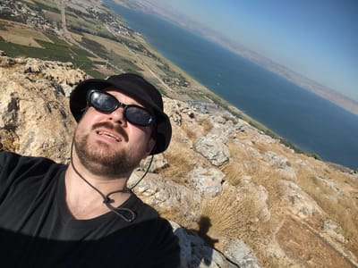 This is a first-person perspective, horizontal, high-angle selfie taken outdoors on a sunny day. The subject, a Caucasian male with a beard, is wearing a black t-shirt and a black bucket hat, and sunglasses. He is positioned in the lower left of the frame, looking up and to his right, with his mouth open as if speaking or laughing. A thin gold chain is visible around his neck. He is standing on a rocky, arid hillside covered with dry grass. In the background, the Sea of Galilee stretches across the frame, with a hazy coastline on the far side. The city of Migdal, Israel, is visible along the coastline, characterized by a road running through it and agricultural fields surrounding it. The sky is clear and blue.