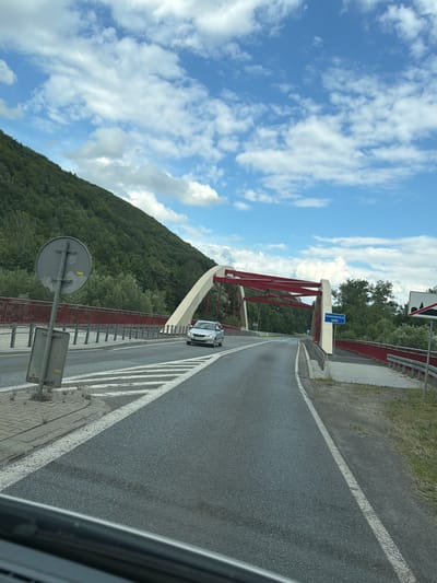 The image captures a view of a road leading onto a bridge. The bridge has a distinctive arching design with red supporting structures. A silver car is driving on the road, crossing the bridge. To the left of the road, there's a large green hill covered in trees. The sky above is blue with scattered white clouds. A blue sign with white text that reads "Kamienica" is visible on the side of the bridge. The scene appears to be a daytime shot in a natural, possibly rural, setting. Given the sign, the location is likely in or near Kamienica, which is a village in the administrative district of Gmina Frycowa, within Nowy Sącz County, Lesser Poland Voivodeship, in southern Poland.
