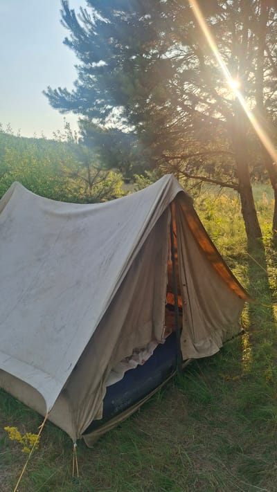 A vintage canvas tent is set up in a grassy, wooded area, likely in the vicinity of Навасвержанскі сельскі Савет, Belarus. The tent is a light tan color and appears to be a traditional A-frame style. Inside the tent, a dark-colored air mattress is visible. The tent is situated among green grass and surrounded by trees, with pine trees prominent in the background. Sunlight streams through the branches of the trees, creating a lens flare effect and illuminating the scene with a warm, golden light. The sky above is a clear, pale blue. The overall impression is one of a serene, rustic outdoor setting.