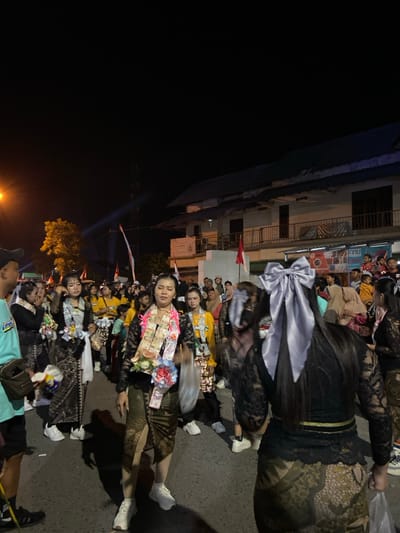 This image depicts a nighttime outdoor gathering or procession on a paved street in the city of Kedungbanteng, Indonesia.

**Scene Overview:** The scene is illuminated by artificial light sources, including an orange streetlight on the far left and spotlights casting a blue beam across the dark sky. Multi-story buildings with balconies and windows line the background on the right. Small red and white Indonesian flags are visible near the buildings.

**People:** Numerous individuals are present, primarily women, forming a crowd. In the foreground, several women wear black long-sleeved lace tops and long patterned skirts, likely batik. Many of these women have garlands draped around their necks, fashioned from Indonesian Rupiah banknotes and artificial flowers. One woman, central in the foreground and facing slightly left, wears such a garland and carries a clear plastic bag. Another woman, in the immediate foreground with her back to the camera, has long dark hair adorned with a large grey satin bow. Other participants and spectators are visible in the background, some wearing yellow shirts. A man in a light blue t-shirt and black cap is partially visible on the far left.

**Actions:** The individuals appear to be standing and moving along the street, suggesting an organized parade or public event.