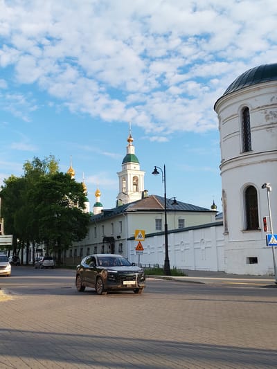 A black SUV drives on a paved street in front of a white wall and several buildings with green and gold domes. The buildings appear to be a religious complex, possibly a monastery or church. The sky is blue with scattered white clouds, suggesting fair weather. The sun is casting long shadows, indicating it is either early morning or late afternoon. The scene is set in Selivanovo, Russia.
Visible text includes a license plate on the SUV which reads "H 648 EC 76". There are also traffic signs. One sign depicts children crossing, another shows a warning symbol with a hill, and a blue sign indicates a pedestrian crossing. A speed limit sign visible on the left shows "40". Another sign shows "↑15KM". A digital display on a pole on the right shows "12:34".