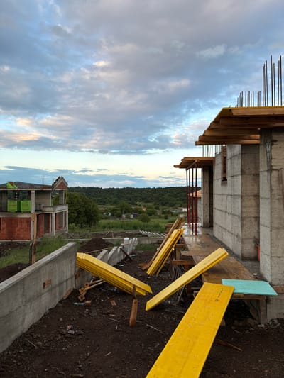 The image shows a construction site located in Cherni Vrah, Bulgaria, with an unfinished concrete structure in the foreground. Exposed rebar extends from the top of the concrete. Yellow wooden planks are scattered nearby on the ground, which appears to be damp soil. In the background, there's a view of a rural landscape with houses and trees. The sky is cloudy with hints of light.
