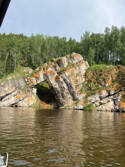 The image shows a natural rock formation arching over a body of water, possibly a river. The rock is layered and shows signs of weathering, with patches of orange lichen and green vegetation growing on its surface. Behind the rock formation, a dense forest of green trees lines a hillside. The water in the foreground is calm, with ripples reflecting the light. The sky is overcast. This natural landmark is located in the vicinity of Martyush, Russia.