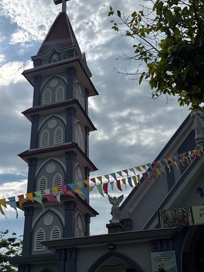 A low-angle, full shot captures the exterior of a church in Suối Tân, Vietnam, on a cloudy day. The church features a tall, grey bell tower with a red-tiled, pointed roof topped by a cross. The tower has several levels, each adorned with arched windows with louvers and decorative trim. Colorful triangular flags are strung across the front of the church. A statue of an angel is positioned on a pedestal above the entrance, and a stained-glass image of two figures is visible on the building. A sign with Vietnamese text is mounted below the stained glass, and another poster with text and a portrait of a man is visible on the lower part of the building. Green foliage from a tree partially obscures the upper right corner of the image. The sky is overcast with dramatic cloud formations.