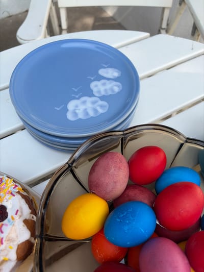 The image depicts a close-up of a collection of Easter items arranged on what appears to be a white outdoor table. To the left, there is a stack of light blue plates with a decorative white floral or leaf pattern. To the right, a clear, multi-tiered serving dish is filled with brightly colored Easter eggs. The eggs are painted in a variety of hues, including red, blue, yellow, orange, and pink. At the bottom right corner of the image, part of a cake or sweet bread, decorated with white frosting and colorful sprinkles, is visible. The scene is set outdoors in Topola Municipality, Serbia.