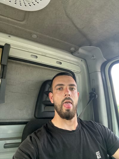 A man is captured in a playful selfie inside what appears to be the cab of a vehicle, likely a van or truck, in Byal Izvor, Bulgaria. He is positioned towards the right side of the frame, looking directly at the camera with his tongue slightly extended. The man has short dark hair and a well-trimmed beard. He is wearing a black long-sleeved shirt, with some white text visible on his right shoulder/arm area, which appears to say "EST OLA".

The interior of the vehicle forms the background, featuring grey, possibly felt or carpeted, lining on the ceiling and walls, along with light-colored plastic panels. A black seatbelt is visible near his left shoulder. In the bottom-left corner, a portion of a white circular object with numerous small perforations, possibly a fan cover or light fixture, can be seen. Through an open window or panel at the top, bright daylight and a glimpse of green foliage are visible, indicating the photograph was taken outdoors during the daytime. The overall mood of the image is casual and lighthearted.