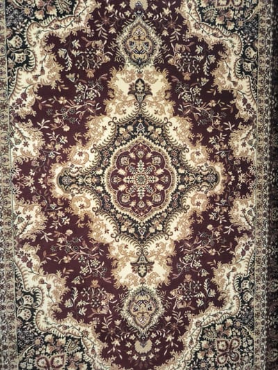 This media file is a close-up, overhead shot of a richly patterned Persian-style rug. The rug features an intricate design with a central medallion and floral motifs. The dominant colors are deep burgundy, cream, black, and gold, creating a luxurious and traditional aesthetic. The texture of the rug appears soft and possibly velvety. There are no people or discernible objects other than the rug itself. The setting is neutral, focusing entirely on the textile. No activity or event is taking place; it is a still image of an object. There are no visible text elements. The lighting is even, suggesting it was taken indoors under artificial light, with no indication of time of day or weather conditions. The image is presented without any specific location cues beyond what can be inferred from the rug's style, which is often associated with Middle Eastern or Central Asian craftsmanship. The overall mood is one of opulent decoration and craftsmanship.