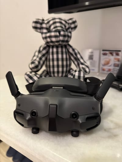 A black and gray plaid teddy bear sits behind a pair of black DJI FPV goggles on a marble countertop. The bear is positioned in the center of the frame, facing forward, with its arms resting on its lap. The DJI FPV goggles are in the foreground, facing the camera, with their antennas extended. The goggles appear to be a new product, possibly for sale or on display. The background is a plain white wall, with a dark shelf or monitor visible at the top of the frame. There is a blurred image of some menus or pictures on the wall in the background. The scene is indoors, with diffused lighting. The location in Ban Phonsavang Neua, Laos, is not depicted in the image.