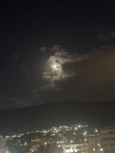 The image is a night-time photograph taken in Tiberias, Israel. The foreground features a portion of the city lights twinkling on a hillside, with buildings and streetlights visible. A dark, wooded area appears between the city lights and the base of the hill. The middle ground is dominated by a dark, silhouetted hillside. The upper portion of the image shows a dark night sky with a bright, full moon partially obscured by clouds. One small star is visible to the left of the moon. The overall atmosphere is serene and nocturnal.