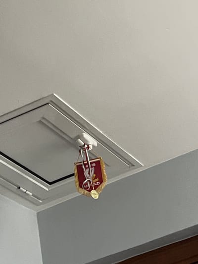 The image shows a white door with a red and gold crest mounted on it. The crest is rectangular with a decorative border and features a design that includes the year "19." The door is located in an interior space with white walls and trim.
