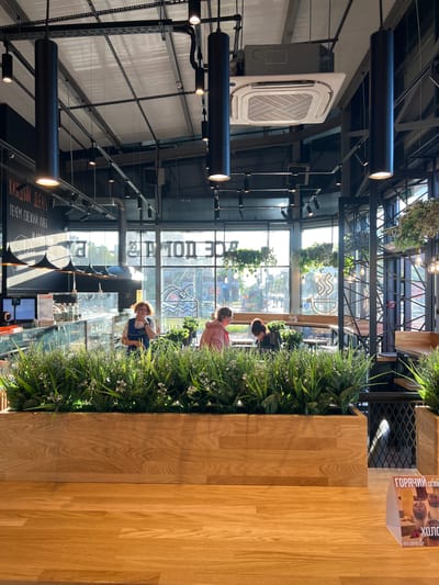 This image depicts the interior of a modern cafe, likely a bakery or coffee shop, with a bright, airy ambiance. The foreground features a large, light-wood planter box filled with lush green artificial foliage, possibly housing small white flowers. Beyond this, a polished wooden counter surface is visible.

In the mid-ground, behind the planter, is a counter area with glass display cases showcasing various products, likely baked goods and drinks. A person wearing a blue apron and glasses is visible behind the counter, appearing to interact with a customer or tend to the display. Further back, two individuals are seated at tables, one wearing a pink hoodie and the other with their back to the camera.

The cafe's ceiling is industrial in design, with exposed beams and a large, square air conditioning unit. Black cylindrical pendant lights hang in clusters, illuminating the space. Large windows dominate the background, offering a view of an outdoor urban setting with trees and buildings. The windows also have large, stylized lettering applied to them, which appears to be in Russian. Hanging plants add a touch of natural greenery to the interior, suspended from the ceiling and structural elements.

On the right side of the foreground, a small triangular sign displays images of drinks, suggesting a promotional offer for hot and cold beverages. The overall aesthetic combines industrial elements with warm wood tones and abundant greenery, creating a welcoming and contemporary atmosphere. The lighting suggests it is daytime, with sunlight streaming through the windows. The image was taken at the Borisova Griva cafe, located in Borisova Griva, Russia.