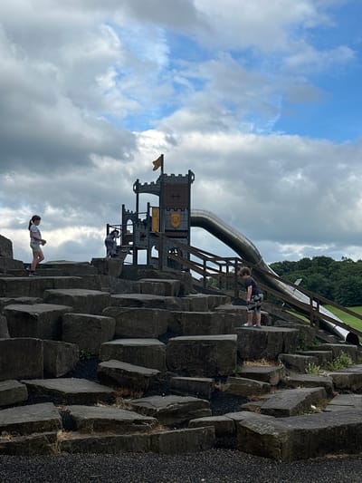 The image depicts a children's adventure playground with a castle-themed climbing structure and a large metal slide. The playground is situated outdoors, with a cloudy sky overhead. In the foreground, a series of large, irregularly shaped dark grey rocks form a stepped climbing area.

There are three children visible. On the left, a young girl with dark hair tied in a ponytail stands facing left, wearing a light blue t-shirt and light grey shorts. In the middle of the frame, another child, whose gender is not clearly discernible due to their clothing (grey outfit), is climbing on the rock structure near the playground equipment. On the right, a boy with curly brown hair is standing on the rocks, looking down, wearing a dark t-shirt with a white pattern on the shorts and blue and white striped flip-flops.

The castle structure is multi-level with a grey and yellow color scheme, featuring turrets, windows, and climbing nets. A yellow flag with a crest flies from one of the turrets. The large, curved metal slide descends from the castle structure. Wooden railings line the path leading to and from the slide.

Beyond the playground, a dense green forest can be seen, with a grassy slope to the right of the slide. The ground around the rocks is covered in dark, coarse gravel. The overall impression is a naturalistic and adventurous play area. The location is identified as a children's play area in the city of Wakefield, United Kingdom, specifically within a park setting given the presence of the playground equipment, rocks, and surrounding greenery.