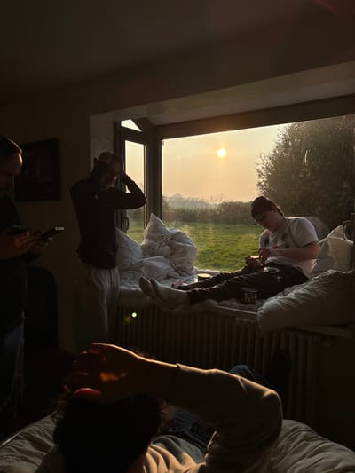 An indoor scene in Bû, France, is depicted from an oblique angle, looking towards a large window that reveals an exterior landscape under a setting or rising sun. The room is dimly lit, with strong backlighting from the window creating silhouettes of the occupants.
Four individuals are present. In the immediate foreground, positioned at the bottom right, a person is reclining, their head and right arm visible. Their right hand is raised to their head, partially illuminated by a direct ray of sunlight. To the left, a second individual stands, facing partially right, engrossed in two smartphones held horizontally in both hands. Next to them, a third individual stands, facing the window, with both hands raised to their head, obscured by shadow. Seated on the window sill to the far right is the fourth individual. This person wears a white t-shirt, dark joggers with a white logo on the left thigh, white socks, glasses, and a dark beanie. They are actively manipulating a small object in their hands. Around this individual on the window sill are several white pillows, a crumpled white duvet or blanket, and a small round container with a white lid, along with another cylindrical object.
Below the window sill, a white radiator is visible. Beyond the window, a bright, low sun is centered in the upper frame, casting an orange-yellow glow across a hazy sky. The outdoor environment consists of a verdant grassy field, with scattered trees and shrubs forming a distant horizon line.
