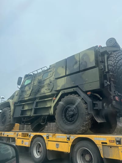 The image shows a military vehicle with camouflage paint loaded onto a yellow transport trailer. The vehicle has a spare tire mounted on top, multiple armored panels, and heavy-duty tires. The trailer has dual wheels on the visible axle. The environment appears to be an overcast day. The location, Kazyli, Russia, is based on a request within the prompt. There are no visible people.
