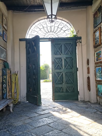 The image depicts an open doorway leading outside, likely a courtyard or garden, bathed in bright sunlight. The doorway features a large, dark green, double wooden door with a decorative lattice pattern on each panel. Above the door is a semi-circular arched window with an intricate wrought-iron grille. To the left of the doorway, a simple wooden bench and a coiled yellow hose are visible. The floor is paved with large, rough-hewn stone tiles, and sunlight casts diagonal shadows across them. On the walls flanking the entrance, several framed artworks and carved wooden masks are displayed. The overall impression is rustic and possibly historic. Without further information, a precise location within Pozzuoli, Italy, cannot be determined.