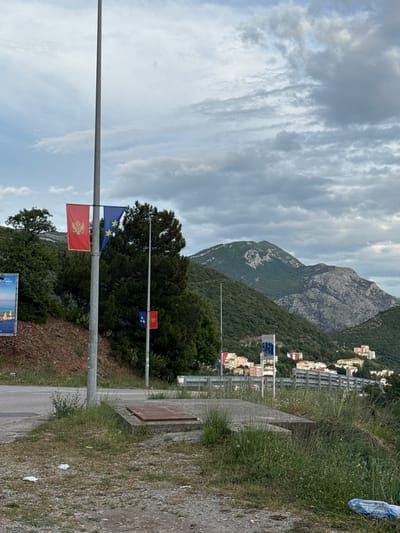 The image shows a roadside scene near Medinski Krš, Montenegro. The foreground features a metal pole displaying the flags of Montenegro and the European Union. Beyond the pole are trees and vegetation. Further in the background is a mountain, with buildings visible on its slopes. The sky is overcast with layered clouds. A paved road runs alongside the grassy shoulder in the lower right corner of the frame.