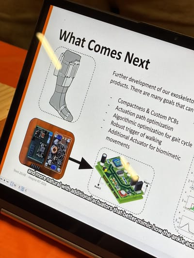 The image shows a screen displaying a presentation slide titled "What Comes Next," which appears to be about the future development of exoskeletons. The slide includes a diagram of a leg exoskeleton and images of circuit boards labeled "Board-1" and "Board-2." Bullet points list goals like compactness, actuation path optimization, algorithmic optimization for gait cycle, robust trigger of walking, and additional actuators for biomimetic movements. Text at the bottom reads "and more natural with additional actuators that better mimic how the ankle act" and "Team 26148 · Imagine RIT 2026," indicating this is a project presented at the Imagine RIT festival held in Rochester, NY.