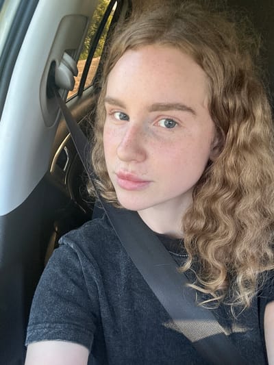 A young woman with wavy blonde hair and fair skin with freckles is seated in the passenger seat of a car. She is wearing a dark grey t-shirt and a black seatbelt is visible across her chest. The interior of the car, including the seatbelt buckle and a portion of the car door, is visible in the foreground. Sunlight streams in from the window, illuminating the scene and casting shadows. The background shows trees and foliage, suggesting an outdoor setting, possibly along a road. The location is identified as Vasil'evo, Russia.