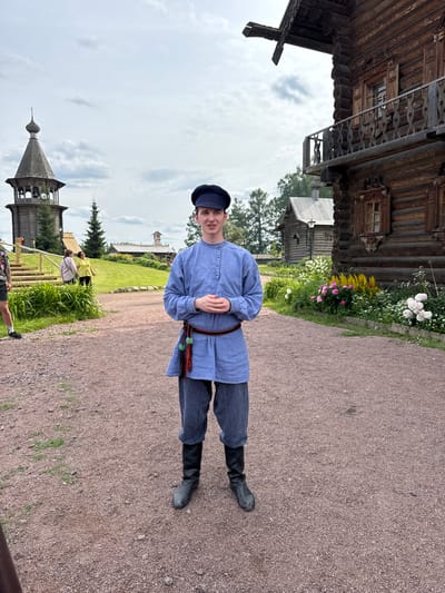 A young man stands in the foreground of an outdoor scene in what appears to be a historical village or park in Nevskii Parkleskhoz, Russia. He is dressed in traditional Russian attire: a blue, long-sleeved shirt with buttons down the front, a dark belt with red and green tassels, and striped blue pants tucked into black boots. He wears a dark flat cap. His hands are clasped in front of him.

Behind him, a path made of gravel leads towards several wooden buildings. To the left, a tall, multi-tiered wooden bell tower with a pointed roof stands against a cloudy sky. Further back, other wooden structures and trees are visible, along with some people walking. To the right, a large, ornate wooden building with a balcony and carved details is prominent, with a flower bed in front of it featuring pink, yellow, and white blossoms. The overall setting suggests an outdoor cultural or historical attraction.