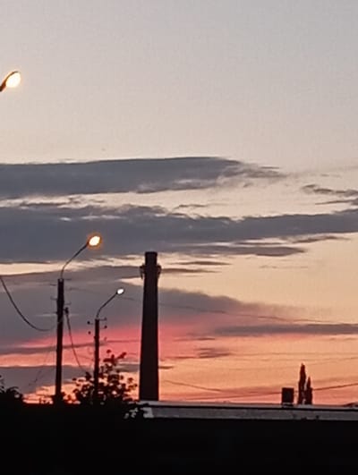 The image shows a silhouetted industrial chimney against a twilight sky with streaks of pink and orange. Several utility poles with lights are visible in the foreground, casting a glow. The scene is likely a part of the industrial area in Yanaul, Russia, given the presence of a prominent chimney and the overall atmospheric conditions. The foreground is dominated by dark, silhouetted structures and vegetation, obscuring finer details.