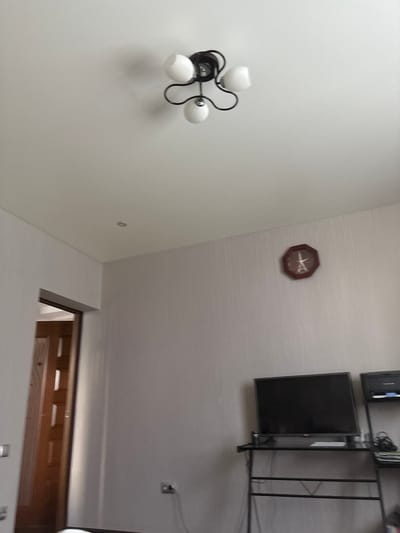 The image shows an interior scene, likely a room in a residential building. A ceiling light fixture with three white globes and a black frame is visible at the top of the image. To the right on the wall, a round, brown clock with a decorative design is mounted. Below the clock, a flat-screen television is placed on a black metal stand. The television is off, displaying a black screen. To the right of the television, another shelf unit is partially visible, holding electronic equipment and cables. A doorway leading to another room is seen on the left side of the image. The walls are painted a light color, and a power outlet with a cord is visible near the floor on the right side of the image. The overall impression is of a living space or a study area. The image does not provide any specific information about the city of Kinel'-Cherkasy, Russia, or any individuals present.