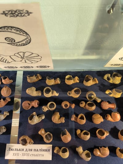 This image depicts an indoor museum exhibit, likely located within the Khotyn Fortress museum in Khotyn, Ukraine. The scene is static, showcasing an array of historical smoking pipes within a glass-enclosed display case.

The primary subjects are approximately 50 to 60 small, individual smoking pipe bowls, meticulously arranged on a dark blue fabric surface. These pipe bowls vary in color from light beige to reddish-brown and dark brown, suggesting different clay types or firing methods. Their forms are diverse, including simple open bowls, some with intricate decorative elements such as fluting, ridges, or sculpted patterns. Many exhibit signs of age and archaeological recovery, including chips, cracks, or minor breakage.

Above the displayed pipes, under the same glass, a rectangular white informational sign is positioned in the lower-left corner. The text on this sign, in Ukrainian, reads "Люльки для паління XVII - XVIII століття," which translates to "Smoking pipes XVII - XVIII century," indicating the artifacts' period. Further up in the display, a light-colored paper sheet with black line drawings is visible, featuring stylized motifs including an eye-like shape with internal patterns and decorative borders. The display case itself consists of clear glass or acrylic, reflecting faint overhead light sources and parts of the room. No people are present, and no actions or interactions are occurring within the scene.