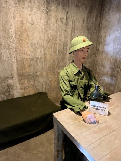 The image captures a historical exhibit inside a dimly lit, narrow underground space, likely a bunker or tunnel. The main subject is a mannequin dressed as a soldier from the People's Army of Vietnam, wearing a light olive green uniform, including a long-sleeved shirt, trousers, and a matching pith helmet with a red star insignia. The mannequin has realistic facial features and light brown skin.
It is posed as if sitting on a lower bunk or wooden platform, with its upper body visible. Its right hand is clenched in a fist, resting on a light wooden surface in front of it, which could be a table or an upper bunk. In its left hand, the mannequin holds a dark green canteen or flask.
The setting features rough, textured walls, varying in shades of brown, grey, and beige, suggesting compacted earth or concrete typical of subterranean structures. A dark green fabric, possibly a tarp or bedroll, is visible beneath the wooden surface.
A white rectangular sign rests on the wooden surface near the mannequin's right hand. The text on the sign is in both Vietnamese and English, stating: "KHÔNG CHẠM VÀO HIỆN VẬT / PLEASE DO NOT TOUCH." This indicates the display is a protected artifact. The scene portrays a static moment, likely depicting a soldier resting in a confined space. Given the location context of Trân Châu, Vietnam, this exhibit most probably represents a scene from the Vietnam War, possibly within a tunnel complex like Củ Chi. The environment is lit artificially, with no external weather or specific time of day discernible.