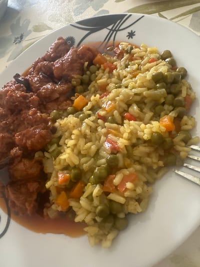 An overhead, close-up shot shows a plate of food featuring a portion of meat in a reddish-brown sauce next to a portion of rice mixed with vegetables. The meat appears to be in bite-sized pieces and is coated in a thick sauce. The rice is a pale yellow color and is mixed with green peas, diced carrots, and small pieces of red bell pepper. A fork with three tines is partially visible on the right side of the plate, resting on the rice. The plate is white with a black and white abstract pattern around the rim. The food is placed on a tablecloth with a floral pattern in muted colors of green, beige, and white. This image was taken in Mainburg, Germany.
