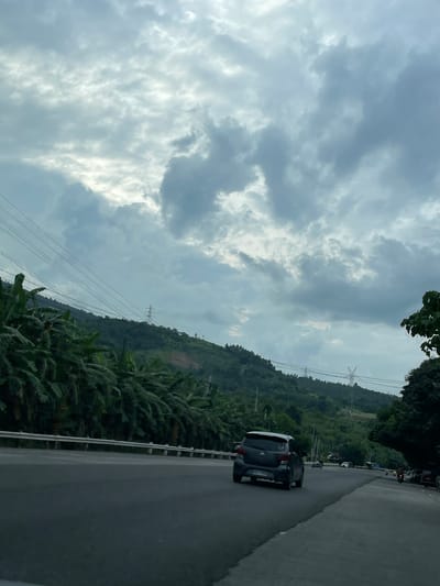 The image depicts a road scene in Santa Cruz, Philippines. A dark-colored car is driving on a paved road. Lush vegetation, including banana trees, lines one side of the road. A mountain or hill is visible in the background, with power lines and transmission towers traversing it. The sky is overcast with heavy clouds. Other vehicles are visible in the distance, suggesting traffic.