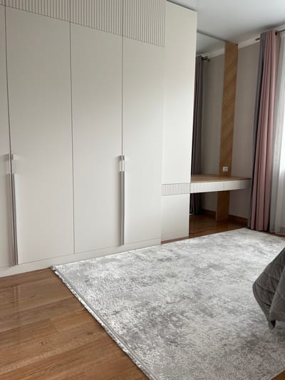 The image shows a bedroom in a modern apartment in Meleuz, Russia. A large, light-colored wardrobe with vertical ribbed detailing at the top occupies the left side of the frame. To the right of the wardrobe, there is a built-in vanity or desk with a wooden countertop and a white, ribbed front panel. Behind the desk, a wooden-paneled accent wall is visible, with two electrical outlets. Sheer pink and gray curtains hang on the right side of the room, framing a window. A light gray rug with a distressed pattern covers a portion of the wooden floor in the foreground, and a corner of a gray quilted bed is visible on the far right.