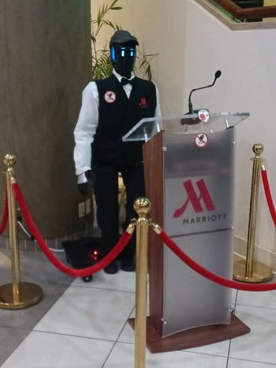 A humanoid robot stands behind a Marriott branded podium with a microphone. The robot wears a gray cap, a white long-sleeved shirt, a black vest, and a black bow tie. Its face appears to be a black screen with blue rectangular "eyes." The scene is demarcated by gold stanchions with red velvet ropes. A plant is visible behind the robot, adding depth to the scene.