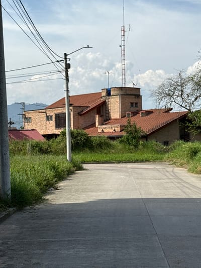 The image depicts an outdoor scene in Pitalito, Colombia, featuring a concrete road leading towards a brick building complex under a partially cloudy sky.

In the foreground, a light gray concrete road, segmented by expansion joints, extends from the bottom of the frame into the midground. Tall green grass and weeds line both edges of the road, more densely on the left. A light-colored utility pole stands vertically on the far left, supporting multiple dark electrical wires and a gray street light fixture.

The midground is dominated by a multi-story building complex constructed primarily of light brown brick, featuring sloped roofs covered with red terracotta tiles. A prominent circular brick tower section rises from the center of the complex, equipped with small windows and topped by a tall lattice communications tower with a red ladder, along with several other antennas and satellite dishes. A blue tarp-like object is partially visible near the top of this tower. Other parts of the building display windows, some fitted with dark security bars. A metal ladder is attached to the side of the circular tower. Dense green foliage and trees are visible around and behind the buildings, with a leafless tree situated on the right midground. Another utility pole with wires is discernible in the left midground.

The background reveals distant mountains or hills under a sky displaying both blue patches and white cumulus clouds. Shadows are cast by the utility pole and other elements, indicating daylight conditions. No individuals are present in the scene, and no overt actions or interactions are observed.