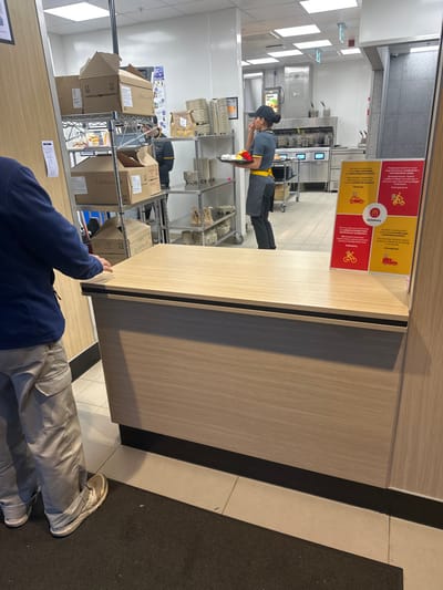 The scene depicts the interior of a McDonald's restaurant located in Chatham, United Kingdom. In the foreground, a partial view of a person is visible from the waist down on the left, wearing light beige cargo pants and white-soled sneakers. Their left arm is resting on a light-wood laminated service counter with a dark horizontal trim. A dark grey mat lies on the tiled floor in front of the counter.
Behind the service counter, a female McDonald's employee stands facing right. She wears a grey and yellow uniform shirt, a dark apron, dark pants, and a dark baseball cap with her hair pulled back. She holds a dark rectangular tray in her left hand, containing a red carton of fries and other wrapped food items.
Behind the employee and to the left, a tall, multi-tiered metal shelving unit is stocked with various cardboard boxes, some open, containing supplies and packaging materials. Another male employee in a similar uniform is partially visible behind these shelves, appearing to interact with the stored items.
The background shows the stainless steel equipment of the kitchen area, including fryers and counters. A sign advertising "Apple Pie" is visible above a station. The walls are white, and the ceiling features recessed fluorescent lighting. To the right of the service counter, a red and yellow "McDelivery" sign displays icons for various delivery methods, with text in multiple languages.