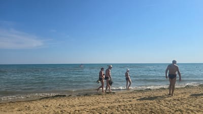 A wide shot shows a sunny day at the beach in San Michele al Tagliamento, Italy. The sand is light brown and textured with some seaweed visible along the waterline. The ocean is a deep blue with gentle waves lapping at the shore. The sky is a clear, bright blue with a faint contrail from an airplane visible.

In the foreground, a man with a tattoo on his back and wearing black swim trunks walks away from the camera towards the water. To his left, a group of three people walks along the shoreline. The first two are a couple, the woman in a black bikini and the man in black swim trunks, carrying a towel and flip-flops. The third person, a woman, is further ahead, wearing a black bikini and a white swim cap.

In the middle ground, a small red boat with a person standing on it is visible on the water. Further out, several other figures can be seen in the water, and some buoys are floating. The horizon line is sharp, separating the water from the sky.