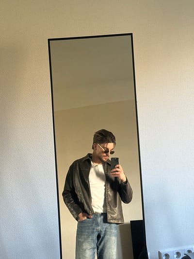 The scene depicts an individual's reflection in a vertical, black-framed full-length mirror, situated indoors in the city of Bertrange, Luxembourg. A male subject, appearing to be an adult, is centered in the reflection, capturing a selfie using a dark-colored smartphone held in his right hand. He is wearing small, oval-shaped sunglasses with thin frames, a dark brown leather jacket, a white t-shirt, and light blue denim jeans. His left hand is positioned in his front left jeans pocket. The background outside and within the mirror's reflection consists of a uniformly textured, light beige wall. In the bottom right corner, outside the mirror's frame, a white electrical outlet with two circular receptacles and a grounding pin is visible on the wall. A dark, indistinct object is partially visible at the very bottom right, adjacent to the mirror. The overall lighting suggests an interior room with indirect illumination.