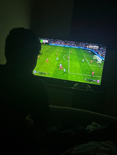 A person is lying down in a darkened room, facing a television. The television displays a soccer video game, with a view of the green field, players, and goal. The scoreboard indicates the game is between teams "CAM" and "BMA," with the score tied at 1-1. Text on the screen also includes "EA SPORTS" and "999.XING." Due to the darkness and angle, specific facial features of the person are indistinguishable. The environment appears to be a residential setting. The location, Welshpool, United Kingdom, cannot be ascertained from the image alone.