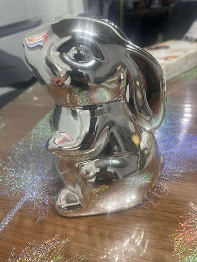 A shiny, metallic, silver-colored ceramic bunny figurine sits on a wooden surface. The bunny is in a seated position, with its head tilted slightly forward and its ears upright. Its surface reflects the surrounding environment, including a person's hand holding a phone, lights, and possibly other objects. The wooden surface is partially covered with a holographic, iridescent material that creates a shimmering, bokeh-like effect. The background is out of focus, showing glimpses of furniture and possibly a book or decorative item. The scene appears to be indoors. The location is not specified in the image or the provided context.