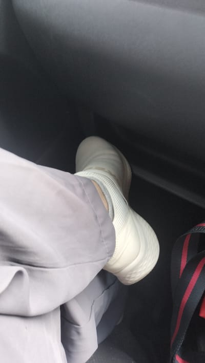 A person's left leg, clad in light gray trousers and a white sock and shoe, is positioned in the foreground of a dark, enclosed space, possibly the interior of a vehicle. The shoe appears to be a slip-on sneaker with a textured sole. To the right of the shoe, a strap with red and black stripes is visible, suggesting the presence of a bag. The background is indistinct and dark, offering little detail about the environment. The scene is devoid of any identifiable landmarks or elements that would place it specifically in Halaban, Indonesia.