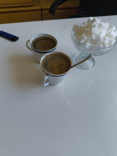 A white table in Hardthausen am Kocher, Germany, holds two white coffee cups filled with dark coffee, a glass dish overflowing with sugar cubes, and a spoon resting in one of the cups. To the left of the cups is a blue object, possibly a pen or lighter. The background shows a wooden surface and a glimpse of a dark chair. The scene is illuminated by artificial light, casting soft shadows on the table.