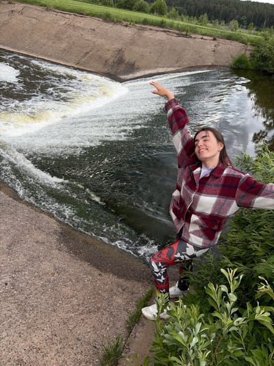 The photograph captures a woman standing near a waterfall in what appears to be a rural or park-like setting. She has light brown hair and is wearing a red and white plaid shirt, black, red, and white leggings, and white sneakers. The woman is smiling and has her arms outstretched as if posing for the photo.
Behind her, a concrete dam or structure directs the flow of water over the edge to create the waterfall. Green grass is visible along the side of the dam. To her right, there are trees and vegetation, some of which she seems to be standing close to.
The image is rotated counter-clockwise. The location is Nizhnii Vishur, Russia.